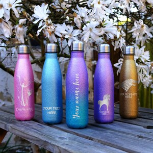 May include: Five glitter-covered water bottles with silver lids are displayed. The bottles are in shades of pink, blue, purple, and gold. Each bottle has a unique design or text, including floral patterns, text that says "Your Text Here", "Your Name Here", a horse illustration, and "Your Shape Here".