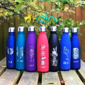 May include: A collection of insulated water bottles in various colors, including blue, purple, and black, each with a silver lid. The bottles feature engraved designs such as a star, owl, and superhero symbols, with names like Isla and Harry.