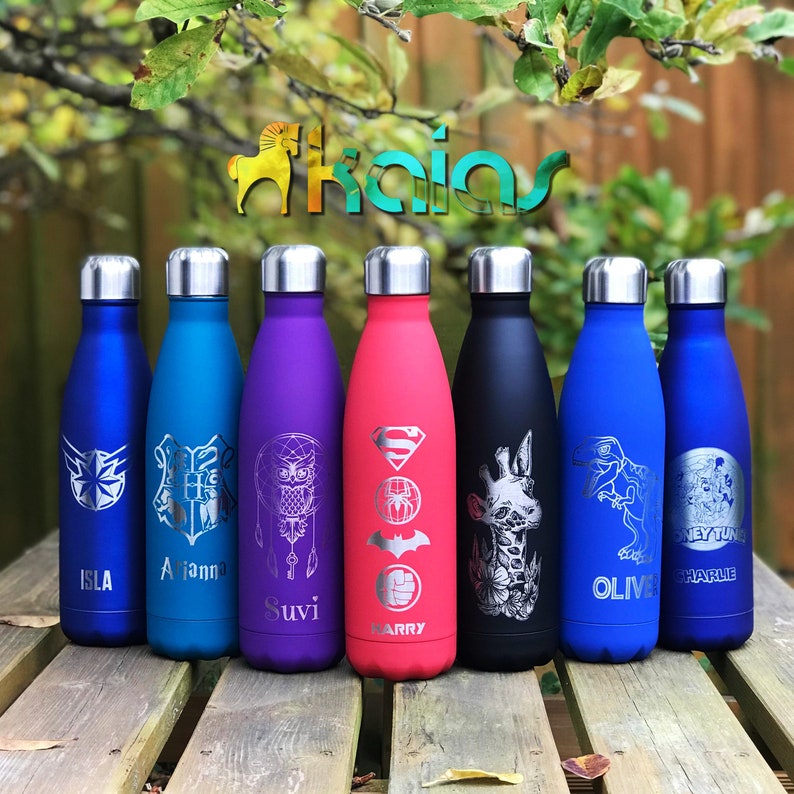 May include: A collection of insulated water bottles in various colors, including blue, purple, and black, each with a silver lid. The bottles feature engraved designs such as a star, owl, and superhero symbols, with names like Isla and Harry.