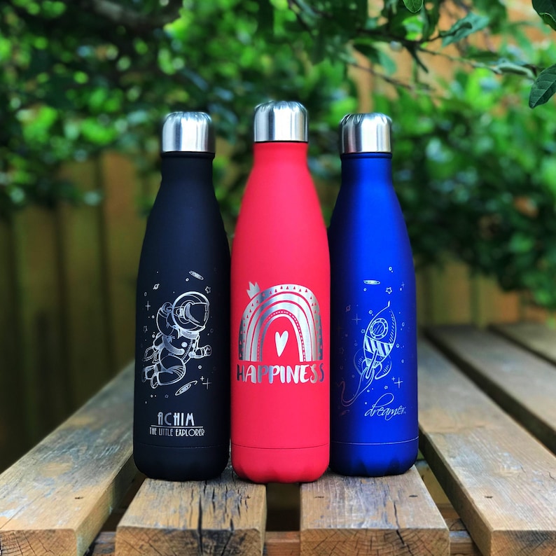 May include: Three insulated water bottles in black, red, and blue, each with a silver lid. The black bottle features an astronaut graphic and the text "ACHIM THE LITTLE EXPLORER." The red bottle says "HAPPINESS." The blue bottle says "dreamer."