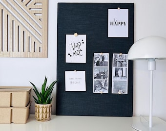 Puede incluir: Un tablero de corcho negro con varios elementos fijados a él, incluyendo una tarjeta blanca con el texto "be HAPPY", una tarjeta blanca con el texto "NUR MUT." en letras negras, una tarjeta blanca con el texto "JUBEL TRUBES HOORAY", y dos fotos en blanco y negro. Una lámpara blanca con una pantalla en forma de hongo se encuentra sobre una superficie blanca delante del tablero de corcho.