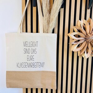 Può includere: Una borsa tote color crema con una base marrone è appesa a una parete a doghe di legno. La borsa presenta il testo tedesco "VIELLEICHT SIND DAS EURE KLASSENARBEITEN!?" ed è abbinata a erba della pampa essiccata e una decorazione a stella di carta.