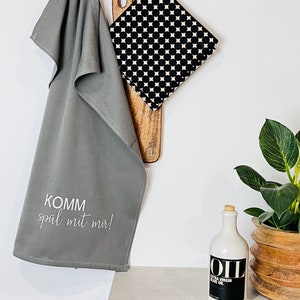 Pode incluir: Uma toalha de cozinha cinza com o texto "KOMMM spiel mit mir!" pendurada em um gancho com um porta-panelas estampado em preto e branco e uma tábua de cortar de madeira. Uma garrafa de cerâmica branca com uma rolha de cortiça e um rótulo preto que diz "OIL EXTRA VIRGIN OLIVE OIL" está em um balcão de madeira ao lado de uma planta em vaso.