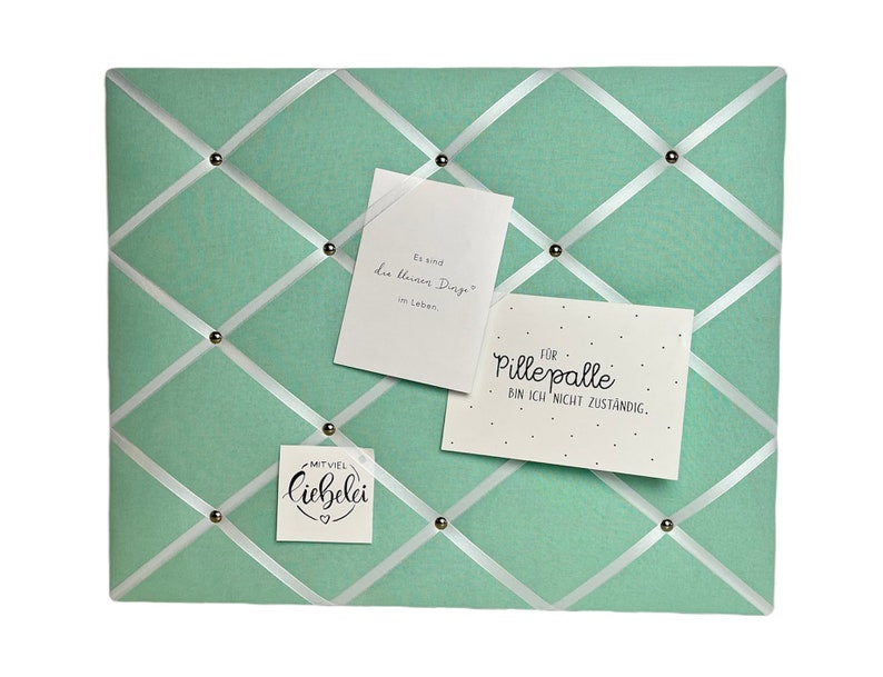 May include: A light green cork board with white ribbon crisscrossing the surface. The board has silver push pins and three white cards with handwritten messages. The cards say "Es sind die leisen Dinge im Leben", "FOR Pille palle BIN ICH NICHT ZUSTANDIG." and "MITVIEL Liebelei ♡".