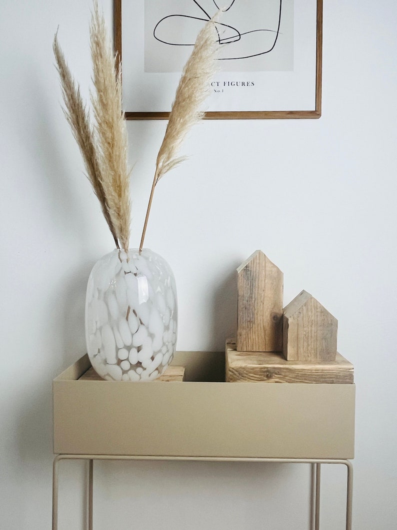 May include: A white ceramic vase with a textured surface filled with dried pampas grass sits on a beige metal plant stand with two wooden houses on top.