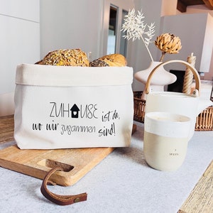 Könnte beinhalten: Ein weißer Canvas-Brotkorb mit einer schwarz-weißen Grafik, auf der "Zuhause ist da, wo wir zusammen sind!" steht. Der Korb ist mit Brötchen gefüllt und steht auf einem Holztisch mit einer weißen Tasse und einem braunen Lederriemen.