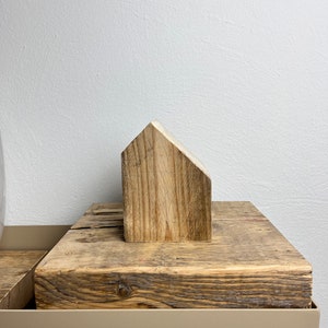 May include: A small wooden house figurine sits on top of a weathered wooden block. The block is on top of a light brown surface.