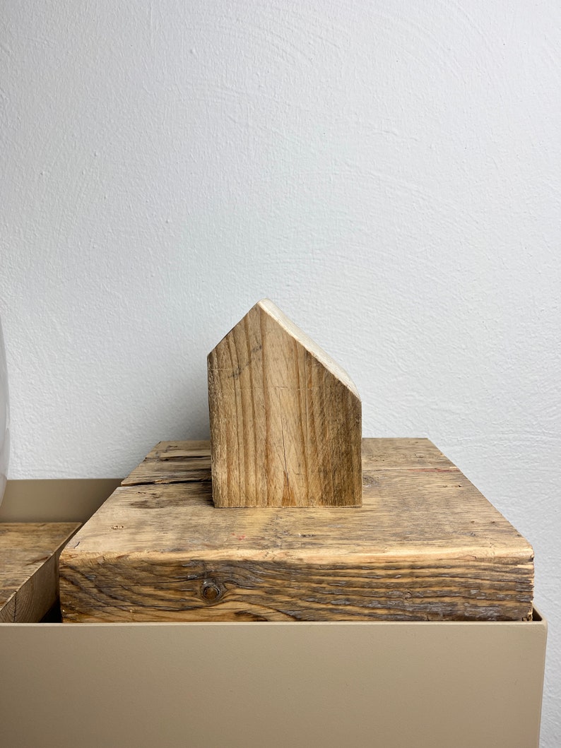 May include: A small wooden house figurine sits on top of a weathered wooden block. The block is on top of a light brown surface.
