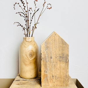 May include: A wooden house-shaped block sits on a rustic wooden shelf with a vase of dried flowers. The vase is made of light wood and has a natural, organic look.
