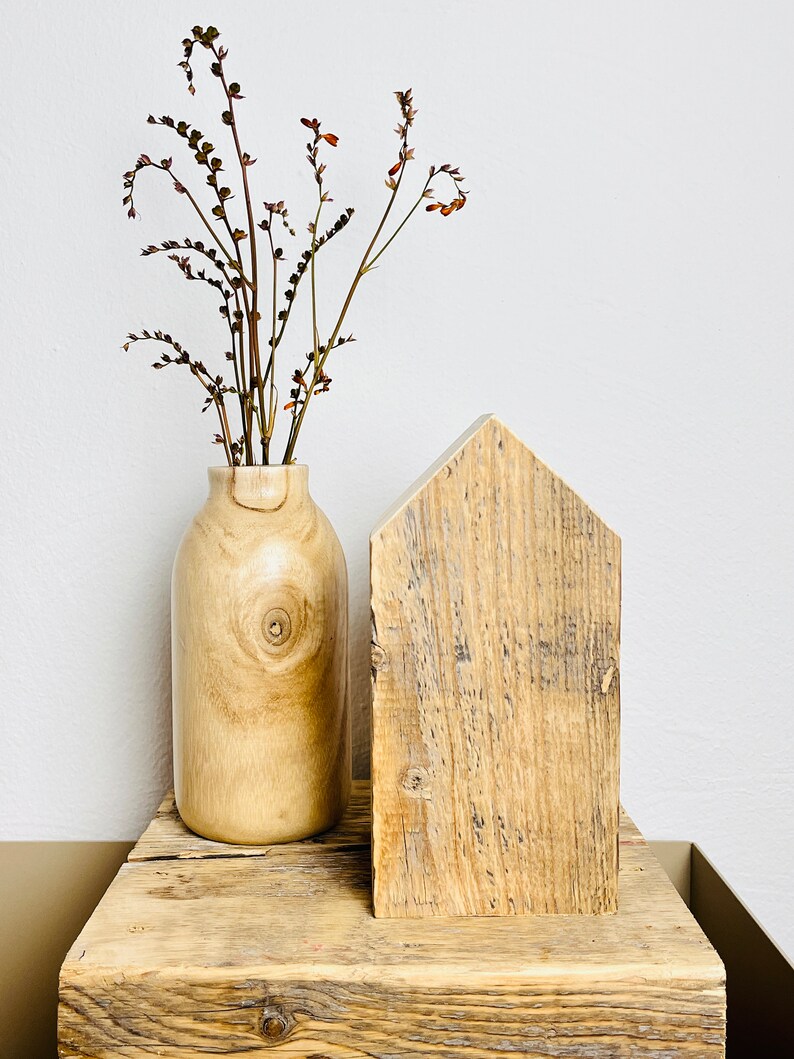 May include: A wooden house-shaped block sits on a rustic wooden shelf with a vase of dried flowers. The vase is made of light wood and has a natural, organic look.