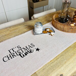 Può includere: Runner da tavolo rosa chiaro con la scritta nera "IT'S CHRISTMAS time" e una stella nera. Su un tavolo di legno, con un vassoio decorativo, una candela e biscotti.