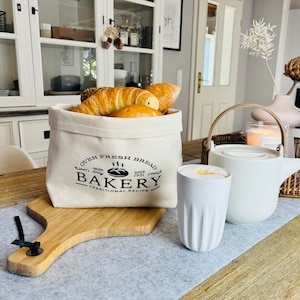 Op de afbeelding: Een witte canvas tas met een zwart logo met de tekst "Oven Fresh Bread Bakery Traditional Recipe" gevuld met broodjes. Een witte keramische theepot met een houten handvat en een witte keramische mok met een latte staan op een houten tafel met een grijze placemat.