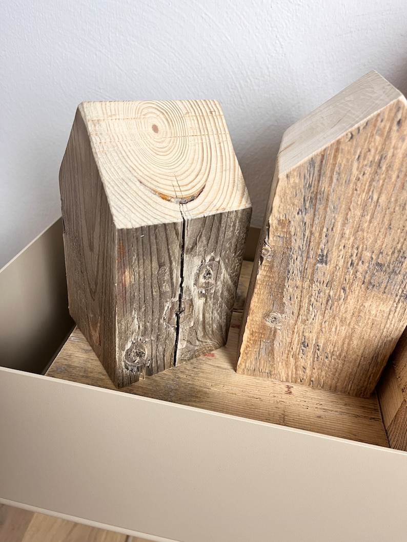 May include: Two wooden house-shaped blocks, one with a light brown finish and the other with a darker brown finish, are displayed in a light beige container.