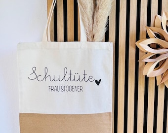 Könnte beinhalten: Cremefarbene Tragetasche mit braunem Boden, mit den schwarzen Schriftzügen "Schultüte" und "FRAU STÖBENER". Ein kleines schwarzes Herz befindet sich neben "Schultüte". Die Tasche hängt an einer Holzlattenwand.