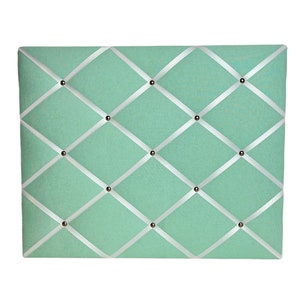 May include: A light green fabric bulletin board with white ribbon crisscrossing the surface. The board is decorated with silver tacks.