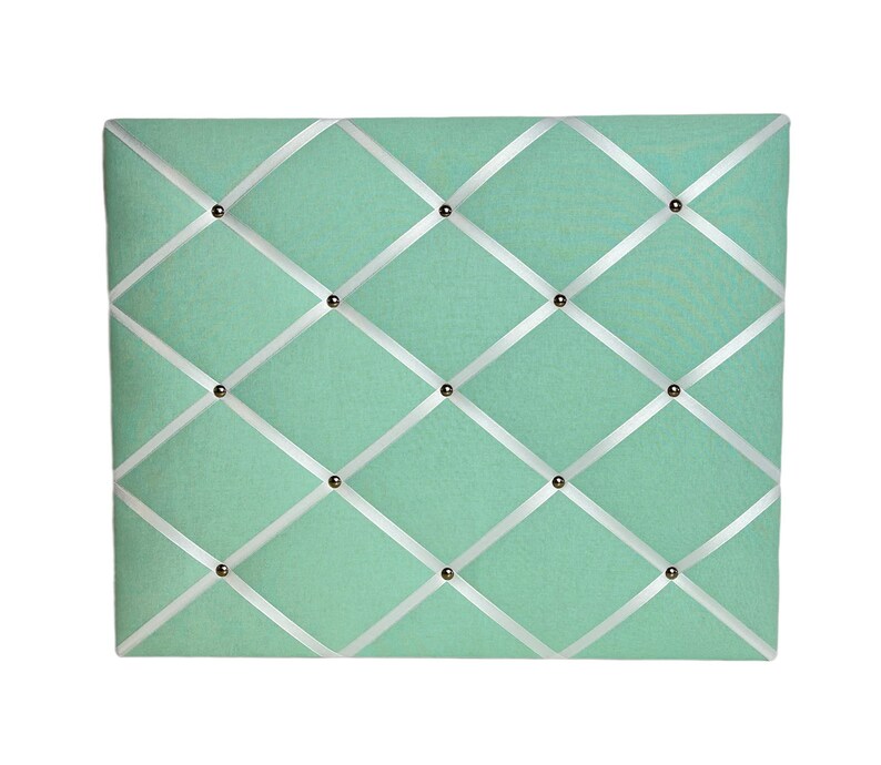 May include: A light green fabric bulletin board with white ribbon crisscrossing the surface. The board is decorated with silver tacks.