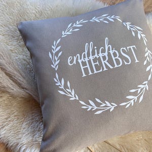 Peut inclure: Un coussin carré de couleur taupe avec le texte allemand "endlich HERBST" en blanc, entouré d'une couronne florale blanche. Le coussin est posé sur une surface moelleuse de couleur crème.