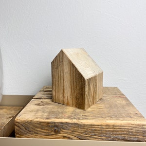 May include: A small wooden house shaped ornament made from natural wood with a light brown color. The ornament is sitting on a wooden surface.