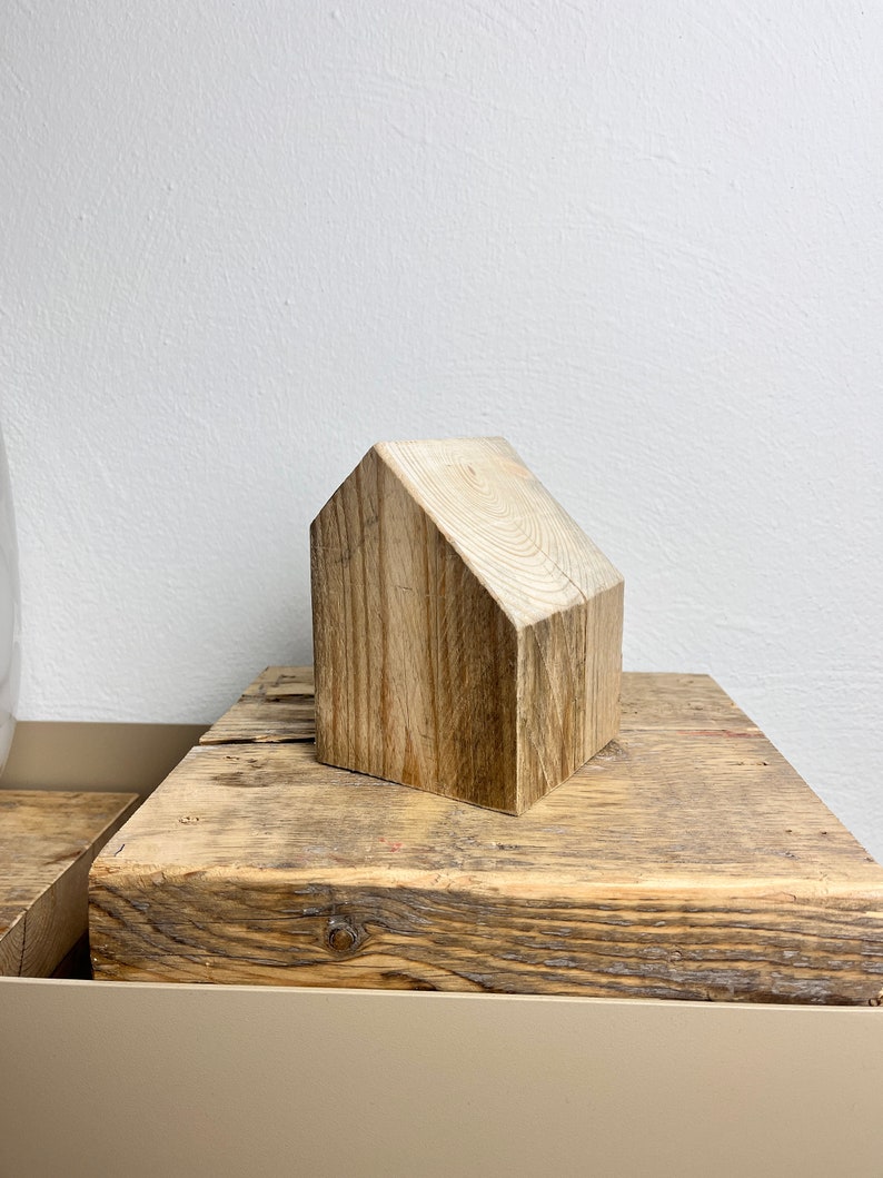 May include: A small wooden house shaped ornament made from natural wood with a light brown color. The ornament is sitting on a wooden surface.