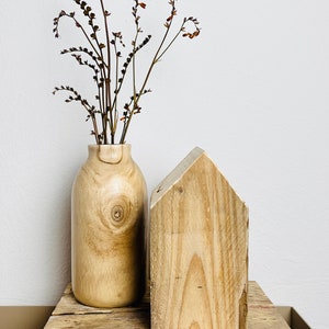 May include: A wooden vase with dried flowers sits on a wooden shelf next to a wooden house-shaped object.