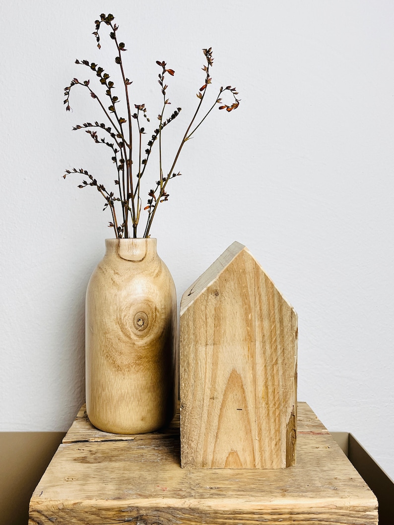 May include: A wooden vase with dried flowers sits on a wooden shelf next to a wooden house-shaped object.