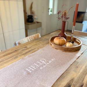 Könnte beinhalten: Ein hellbrauner Tischläufer aus Stoff mit dem weißen Aufdruck "HELLO autumn" in einer Kombination aus Block- und Schreibschrift. Auf dem Läufer steht ein rundes Holztablett mit zwei kleinen orangefarbenen Kürbissen, einer dunkelroten Vase und einer Kerze.