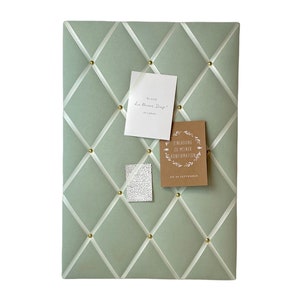 Könnte beinhalten: Eine salbeigrüne Pinnwand mit weißem Band, das sich über die Oberfläche kreuzt. Die Pinnwand ist mit goldenen Stecknadeln bedeckt und hat drei Karten, die daran befestigt sind. Eine Karte ist weiß mit dem Text "Es sind die kleinen Dinge" und eine andere Karte ist braun mit dem Text "Einladung zu meiner Konfirmation am 12. September".