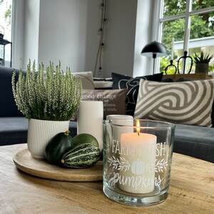 Könnte beinhalten: Eine brennende Kerze in einem Glas mit dem Text "Farm Fresh Pumpkins" steht auf einem Holztablett mit dekorativen Kürbissen und einer Topfpflanze. Die Szene spielt sich auf einem Holztisch in einem Wohnzimmer ab.