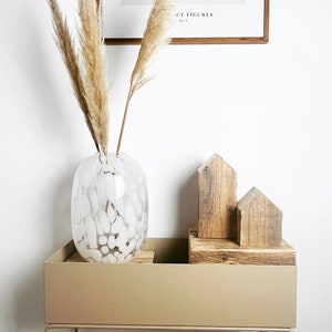 May include: A white ceramic vase with a textured surface filled with dried pampas grass sits on a beige planter with a metal stand. Two wooden houses are arranged on top of the planter.