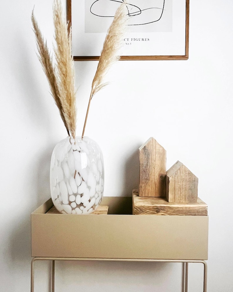 May include: A white ceramic vase with a textured surface filled with dried pampas grass sits on a beige planter with a metal stand. Two wooden houses are arranged on top of the planter.