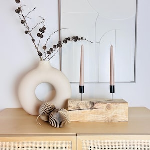 Può includere: Un'arrangiamento decorativo per interni dai toni neutri, con un vaso beige con apertura circolare, due candele rosa in supporti neri e un portacandele in legno. Ornamenti decorativi in carta e un'opera d'arte astratta incorniciata completano la scena. Un ramo con piccole pigne marroni è nel vaso.