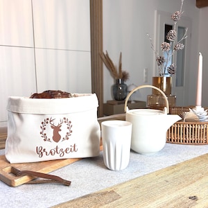 Op de afbeelding: Witte canvas broodzak met een bruin hert silhouet en de tekst "brotzeit" in bruine letters. De zak ligt op een houten tafel met een witte keramische mok en een witte theepot.