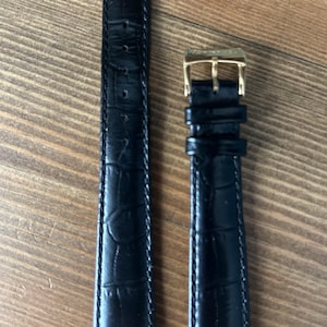 Può includere: Cinturino per orologio in pelle nera con motivo a coccodrillo. Il cinturino ha una fibbia color oro. Il cinturino dell'orologio è mostrato su una superficie di legno.