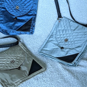May include: Three quilted crossbody bags in shades of blue and silver-grey. Each bag features a flap closure with a decorative button and a black leather strap. The bags have a geometric quilted pattern and a black accent.