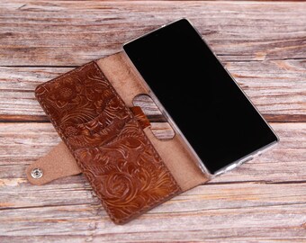 May include: A brown leather phone case with a floral embossed design. The case has a flap that folds over the phone and snaps closed. The phone is not visible in the image.