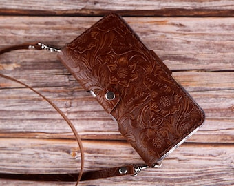 May include: A brown leather phone case with a floral embossed design and a long brown strap. The case is lying on a wooden surface.