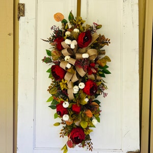 May include: A burgundy and green fall door swag with a burlap bow and white cotton balls. The swag is hanging on a white door.