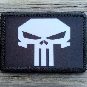May include: A rectangular black patch with a white skull design. The skull features angular eye sockets, a nose cavity, and vertical teeth. The patch has a black border with stitched edges.