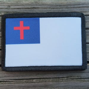 May include: Rectangular patch featuring a blue square with a red cross in the upper left corner, and a white background. The patch has a black border and is displayed on a wooden surface.
