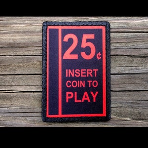 May include: A rectangular patch with a black background and red text. The text reads "25¢ INSERT COIN TO PLAY". The patch has a red border and is likely made of fabric.