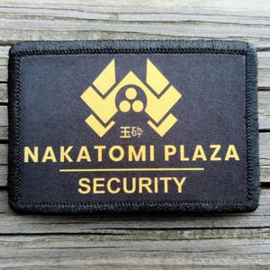 May include: Black rectangular patch with gold text and a stylized emblem. The text reads "NAKATOMI PLAZA" above "SECURITY". The patch has a black border and is set against a wood grain background.