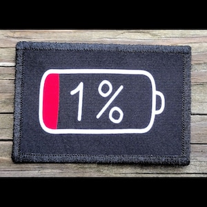 May include: Black rectangular patch with a white outline of a battery showing 1% charge. The battery's red indicator is on the left side. The number "1%" is written in white inside the battery outline. The patch has a black stitched border.