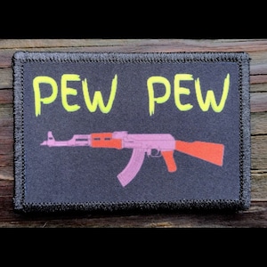 May include: A rectangular patch with the words "PEW PEW" in bright yellow letters above a pink and orange AK-47 rifle design. The patch has a black border and is set against a dark background.