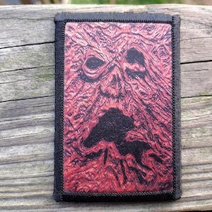 May include: A rectangular patch with a black border features a red and black image of a face with a gaping mouth. The face has a textured, wood-like appearance. The patch is set against a weathered wooden surface.