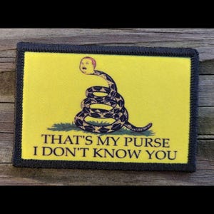 May include: A rectangular patch with a yellow background and a black border. The patch features a coiled snake with a human head and the text "THAT'S MY PURSE I DON'T KNOW YOU" in black letters. The snake is black and yellow.