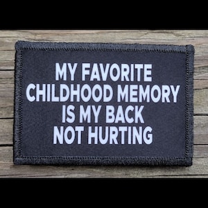 May include: Black rectangular patch with white text that reads "MY FAVORITE CHILDHOOD MEMORY IS MY BACK NOT HURTING." The patch has a black border with stitched edges. The background is a wooden surface.