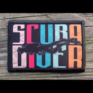 May include: A rectangular patch with the word "SCUBA DIVER" in large, distressed, colorful letters. A black silhouette of a scuba diver is centered on the patch. The patch has a black border and is set against a wooden background.