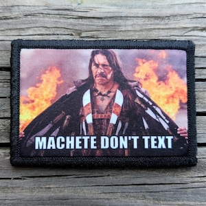 May include: A rectangular patch with a black border features an image of a man with long dark hair, standing in front of flames. The text "MACHETE DON'T TEXT" is printed in white across the bottom of the patch.