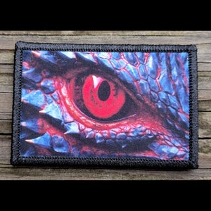 May include: A rectangular patch featuring a close-up of a dragon's eye. The eye is a vibrant red, set against a backdrop of blue and purple scales. The patch has a black border and a detailed, textured appearance.