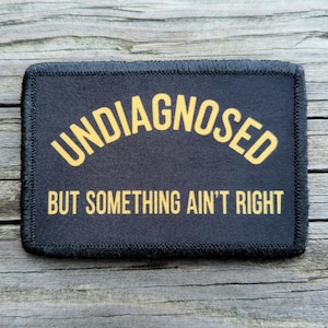 May include: A rectangular black patch with a stitched border featuring the yellow text UNDIAGNOSED in an arched formation above the straight yellow text BUT SOMETHING AIN'T RIGHT. The patch has a clean finish and clear, bold lettering throughout.